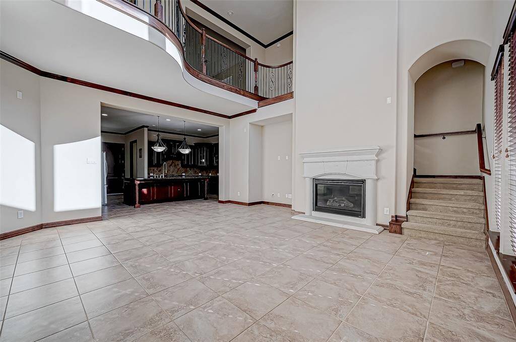 This spacious living area features a high ceiling and an elegant staircase. It includes tile flooring, a modern fireplace, and large windows for natural light. The open layout leads to a kitchen with dark wood cabinets and a stylish island.