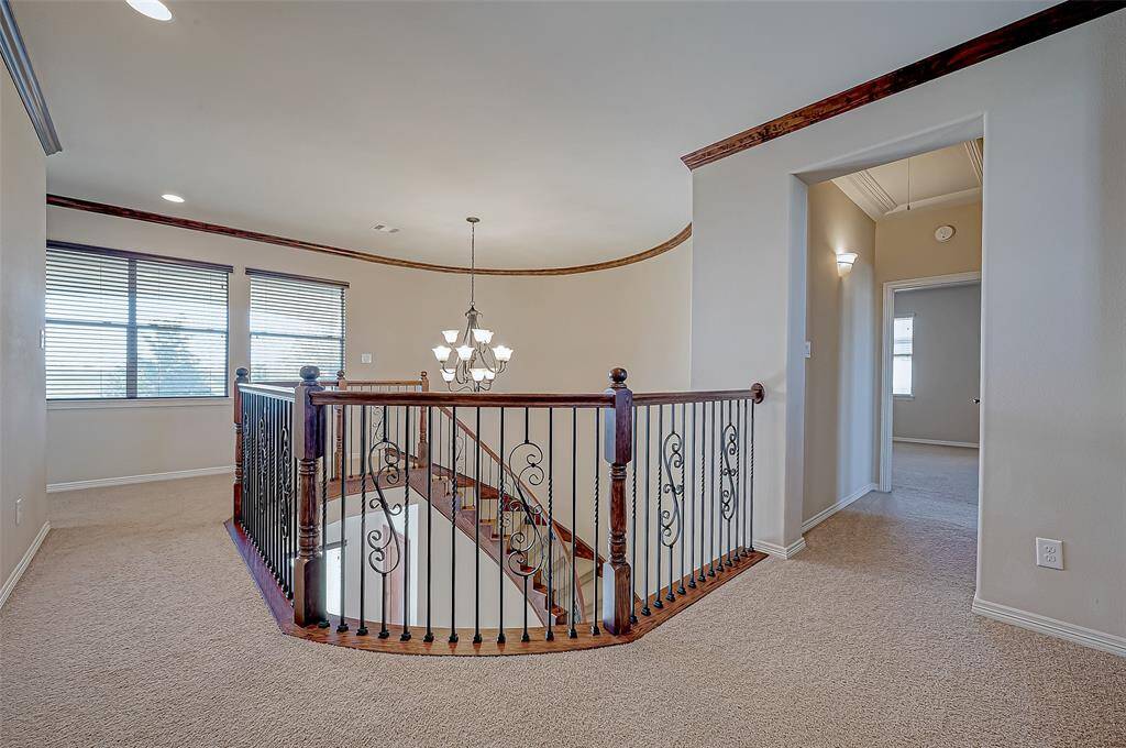 This photo showcases an upstairs landing area with a curved wooden and wrought iron railing overlooking a staircase. The space is carpeted, features recessed lighting, and a decorative chandelier. There are large windows offering natural light, and an entrance to a room on the right.