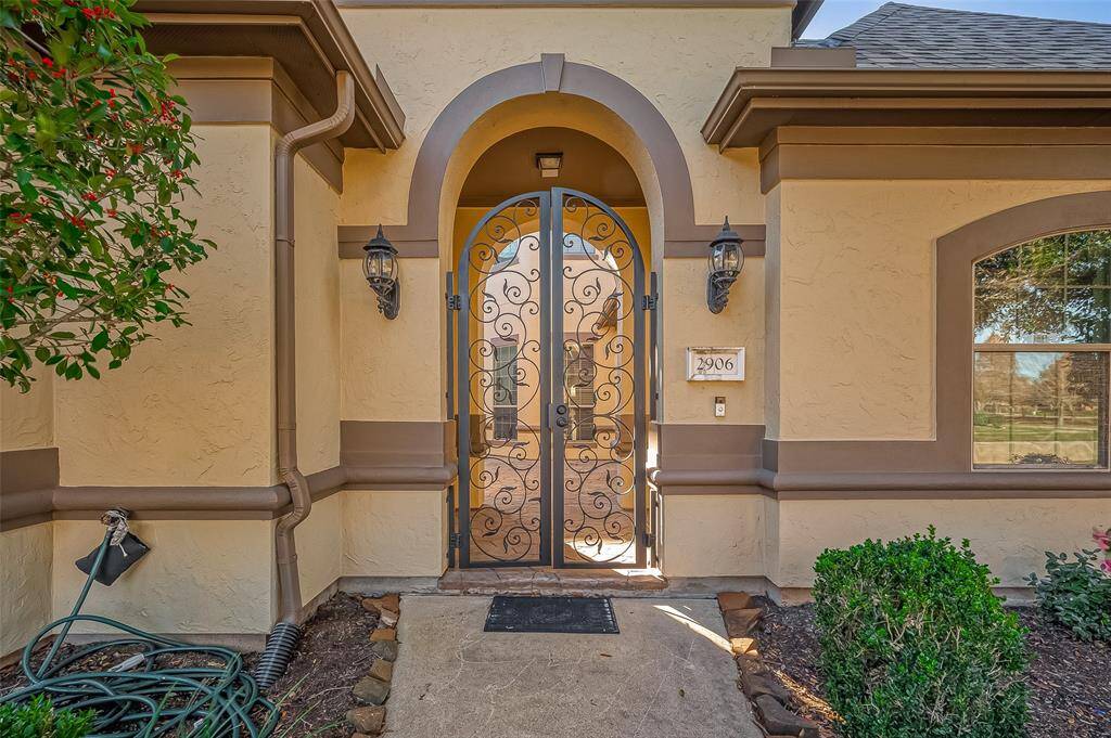 This photo shows an elegant front entrance featuring a decorative wrought iron gate set within an arched doorway. The exterior walls are a warm, earthy tone with contrasting trim, and there are tasteful outdoor light fixtures on either side of the door. Lush greenery and a well-maintained garden border the pathway, adding to the inviting curb appeal.