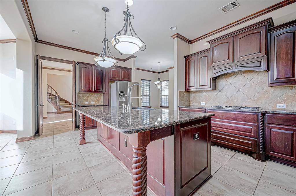 This spacious kitchen features elegant dark wood cabinetry, a large central island with a granite countertop, and modern stainless steel appliances. The tile flooring adds a touch of sophistication, while pendant lighting provides a warm ambiance. An open layout connects to adjacent living spaces.