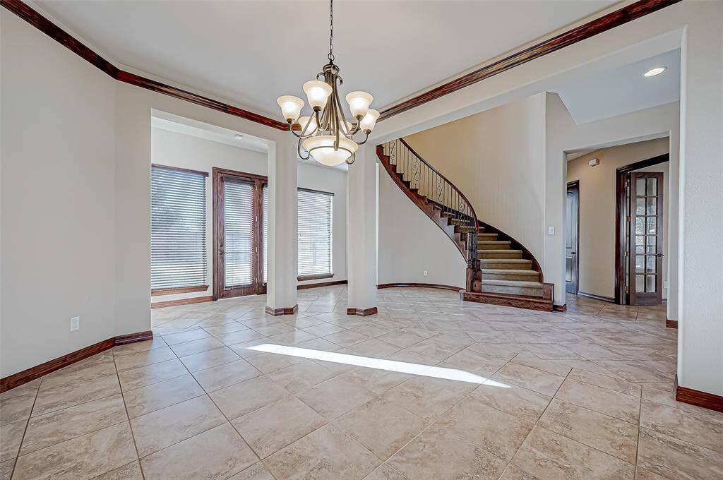 This bright entryway features elegant tile flooring, a grand curved staircase with wood accents, and a stylish chandelier. French doors lead to a sunlit room with large windows, creating an inviting and open ambiance.