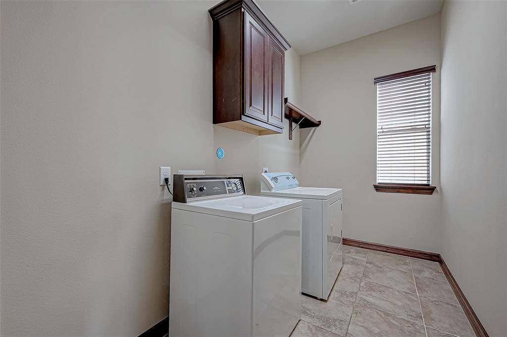 This photo shows a well-lit laundry room with a washer and dryer, tile flooring, a window with blinds, and overhead cabinetry for storage.