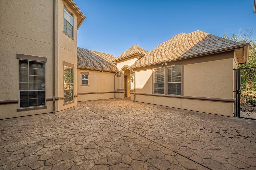 This photo showcases a spacious, paved courtyard with a textured stone-like surface. The exterior features a stucco finish with multiple large windows, creating a bright and inviting space. The architecture includes a peaked roof and a decorative archway leading to the entrance, providing a blend of elegance and functionality.