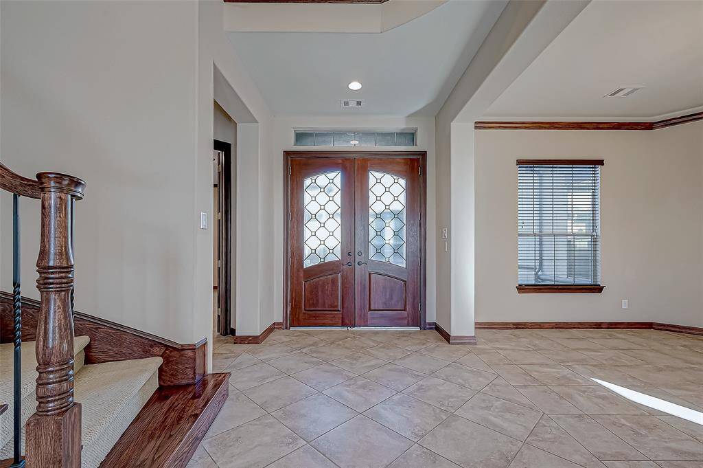 This welcoming entryway features elegant double doors with decorative glass, tile flooring, and a staircase with rich wood accents. Natural light filters through a window with blinds, highlighting the detailed crown molding.