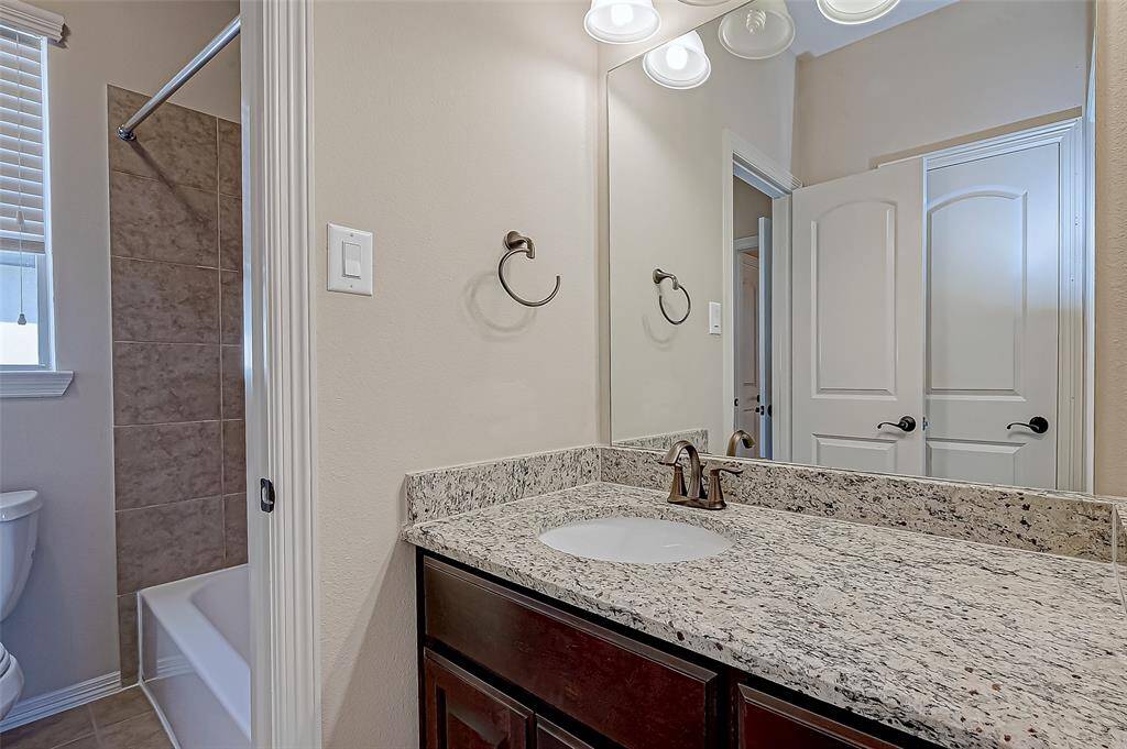 This bathroom features a sleek granite countertop with an undermount sink, a dark wood vanity, and modern fixtures. It includes a tiled shower and bathtub combination, with natural light filtering through a window with blinds. The space is accented by neutral tones and a large mirror.