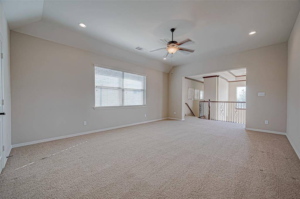 This photo shows a spacious, carpeted room with neutral walls, recessed lighting, and a ceiling fan. There's a large window for natural light and an open view to a hallway or additional area with railing.