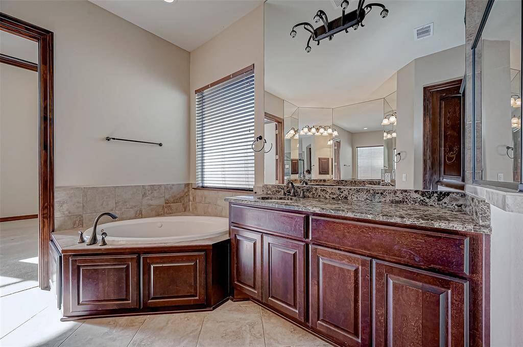 This bathroom features a luxurious corner bathtub with elegant tile surround, a spacious granite countertop with dark wood cabinetry, and a large mirror enhancing the room's brightness. The lighting fixtures add a touch of sophistication to the space.