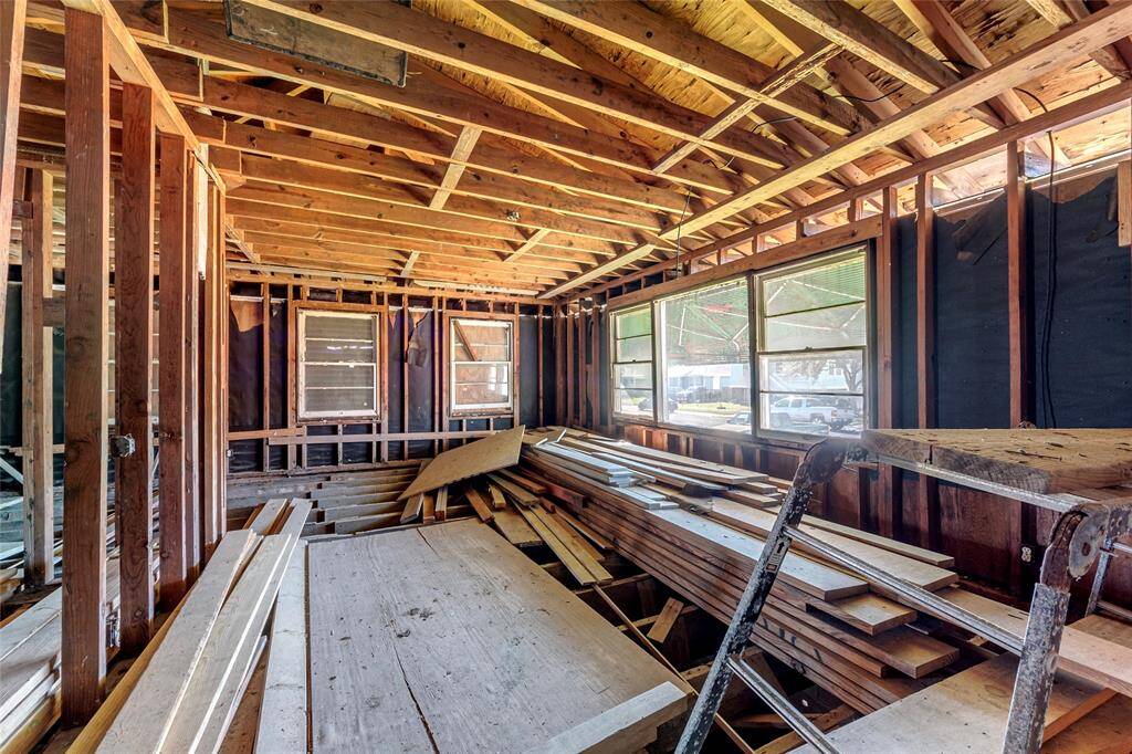 This photo shows a room under construction or renovation, featuring exposed wooden framing and unfinished walls. There are large windows letting in natural light and stacks of lumber on the floor, indicating work in progress.