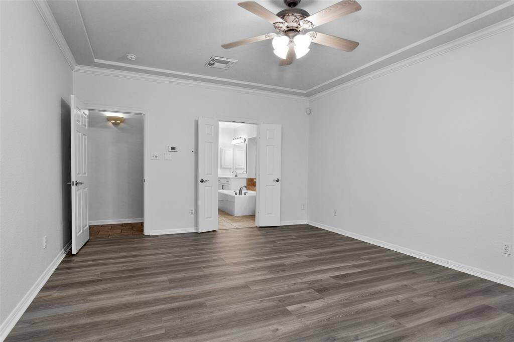 This room features light gray walls, wood-look flooring, and a ceiling fan with lights. It has a doorway leading to the bathroom with a large bathtub and 2 sink areas.