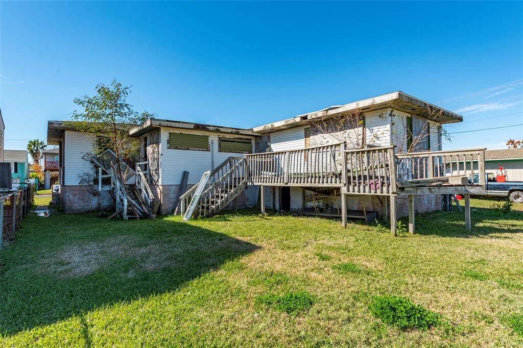 This photo shows a modest home with a raised wooden deck and stairs, set on a grassy lot. The siding is white, and the building appears to need some maintenance. The property is fenced, and there's ample outdoor space.