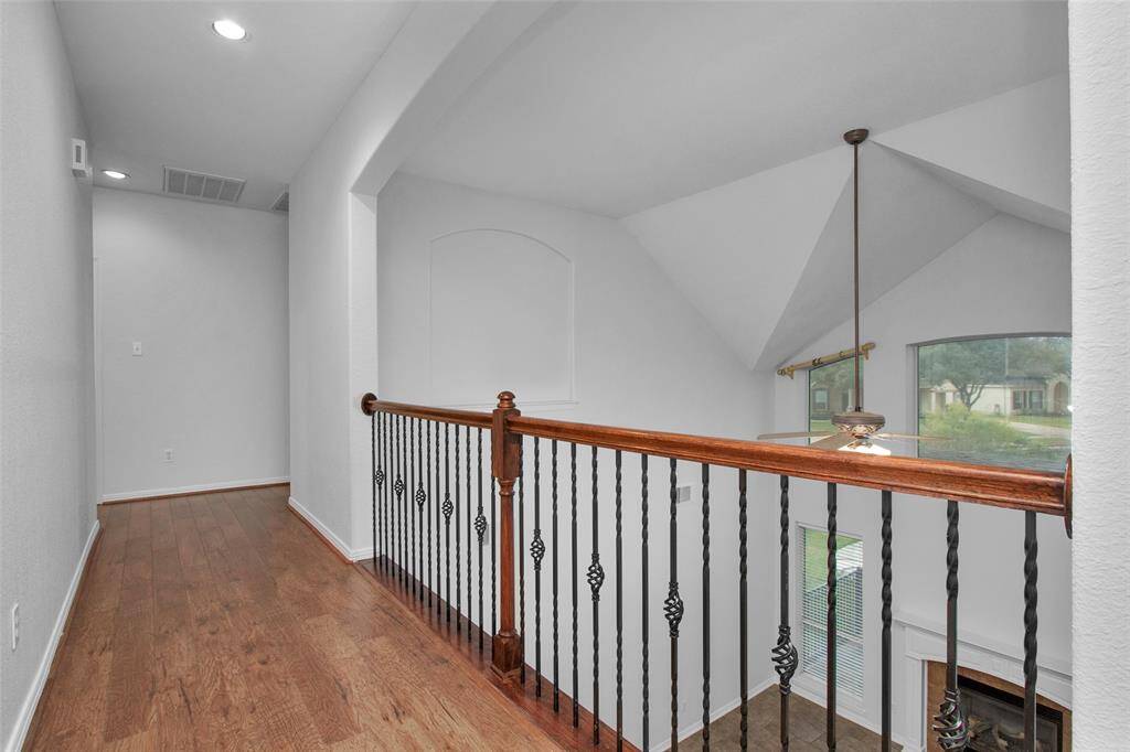The hallway with wood flooring and a decorative iron railing overlooking a bright, vaulted living area. Large windows provide ample natural light, and there’s a view of a ceiling fan and fireplace below.