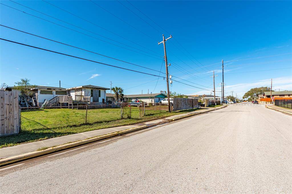 A quiet residential street with single-story homes, a clear blue sky, and ample front yards. The area appears spacious with utility poles lining the street.