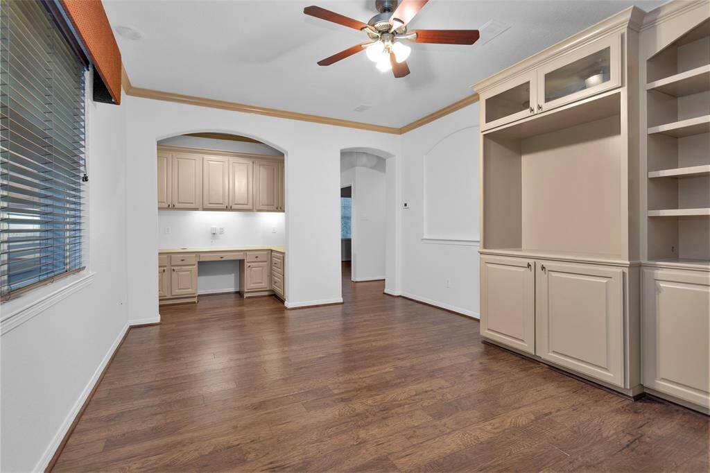 This room features hardwood floors, built-in shelving, and a ceiling fan. It includes a cozy nook with cabinetry, ideal for a home office or study area. Large windows bring in natural light, enhancing the open and airy feel.