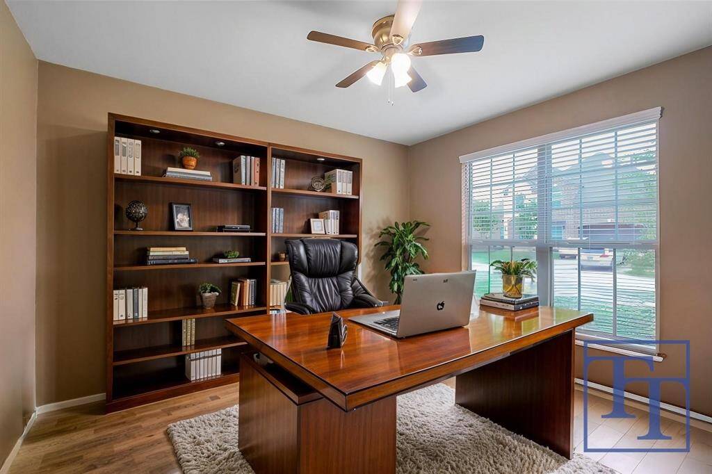 Step into this home office featuring elegant shelving, a spacious desk, and abundant natural light from the large window overlooking a serene neighborhood. The warm tones and sleek design create a productive and inviting workspace.