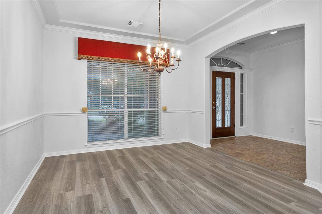This photo showcases a bright, open dining area with elegant light wood flooring, a classic chandelier, and a large window with a red valance. The space flows into an entryway with a decorative glass front door and tile flooring, offering a welcoming atmosphere.