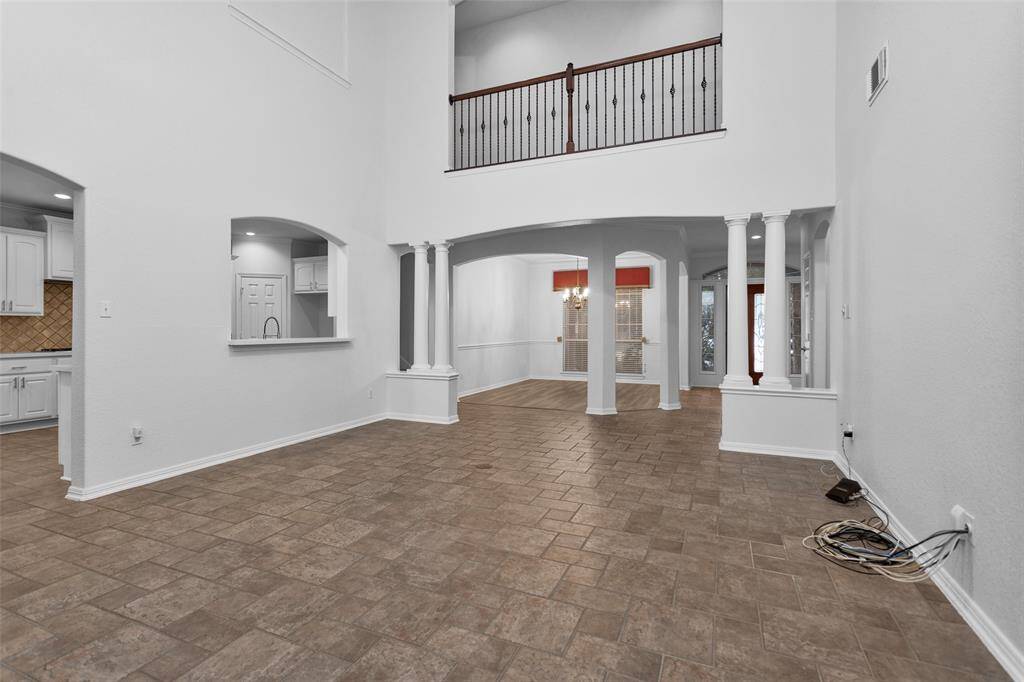This spacious living area features high ceilings and an open layout with tiled flooring. It includes a view of the kitchen with a pass-through window. The room feels bright and airy, enhanced by white walls and architectural columns.