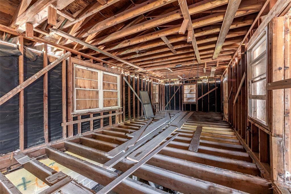 This photo shows a room under renovation or construction, featuring exposed wooden framing, rafters, and missing floorboards. Large windows provide natural light, and the space is ready for customization.