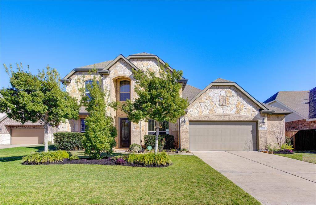 This is a charming two-story stone and brick home with a spacious front yard and a well-maintained driveway leading to a two-car garage. The house features elegant arched windows and a welcoming entrance, surrounded by mature trees and landscaped greenery.