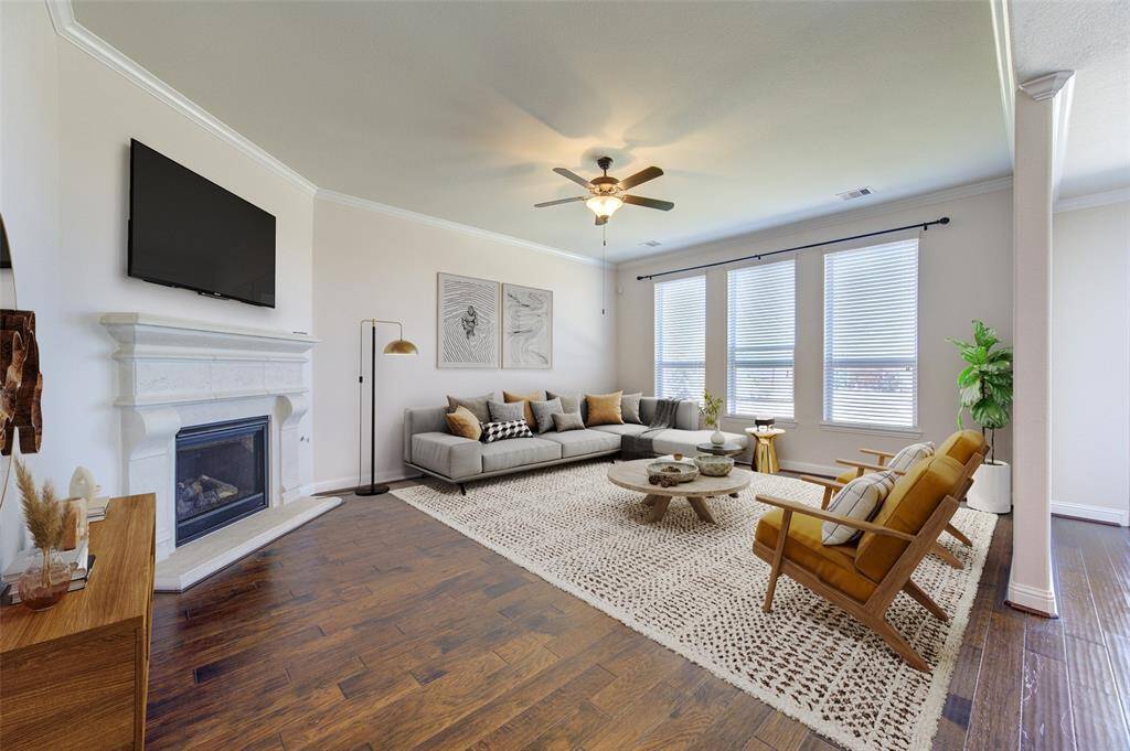 This inviting staged living room features a cozy fireplace with a mounted TV, elegant hardwood flooring, and ample natural light from large windows. The space is tastefully furnished with a modern sectional sofa, stylish armchairs, and a chic coffee table, creating a warm and welcoming atmosphere.