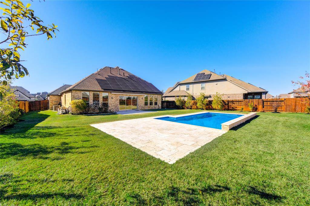 This photo highlights a spacious backyard with a meticulously maintained lawn and a large in-ground pool surrounded by a beautiful stone patio covered area with travertine pavers, offering versatile use—perfect for placing lounge chairs or creating unique spaces to suit your needs. It can be covered or left open, depending on your preferences. .