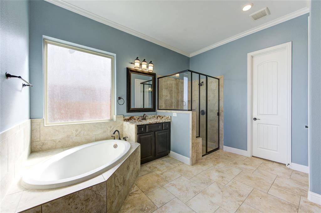 This bathroom features a spacious layout with a large soaking tub, a glass-enclosed shower, and a stylish vanity with a framed mirror. Natural light fills the room through a frosted window, and the walls are painted in a calming blue tone. The floor and tub surround are tiled, adding elegance to the space.