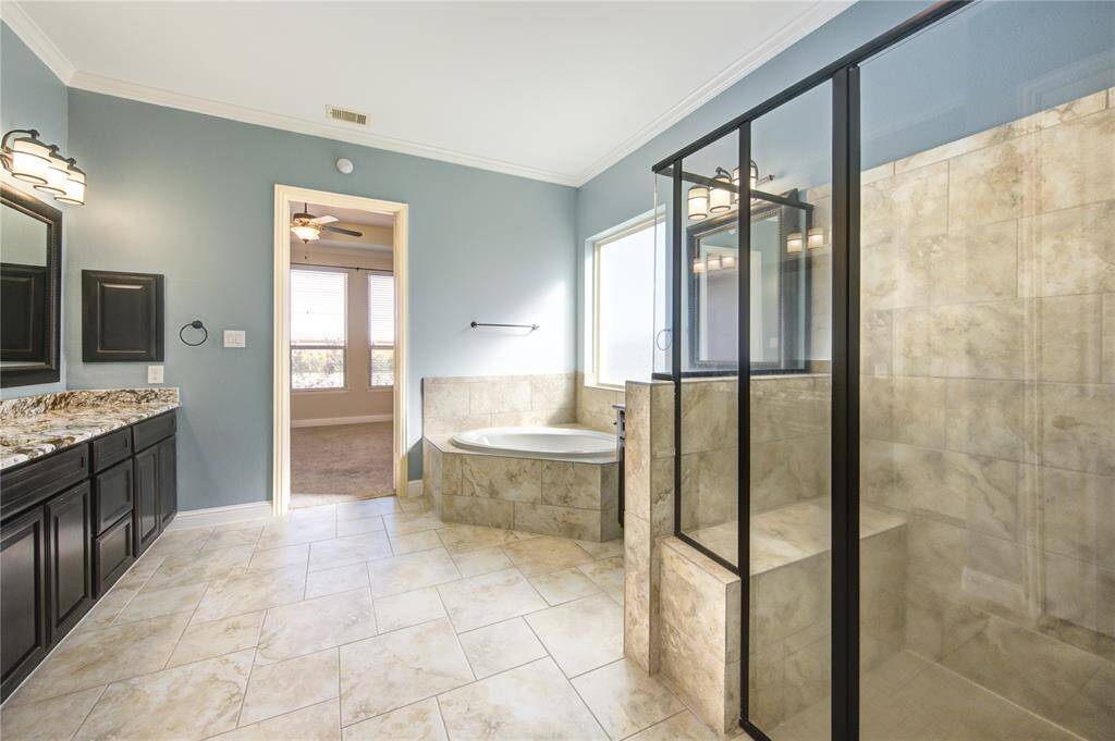 This spacious bathroom features a large walk-in shower with glass doors, a luxurious bathtub, double sinks with granite countertops, and ample cabinet storage. The room is well-lit with natural light from a large window, creating a bright and inviting atmosphere.
