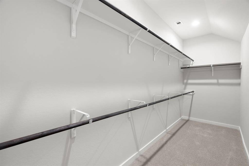 spacious walk-in closet with high ceilings and plush carpet. Warm paint tones, built-in shelving, and dark finishes create a contemporary and functional retreat. Sample photo of completed home with similar floor plan. Actual colors and selections may vary.