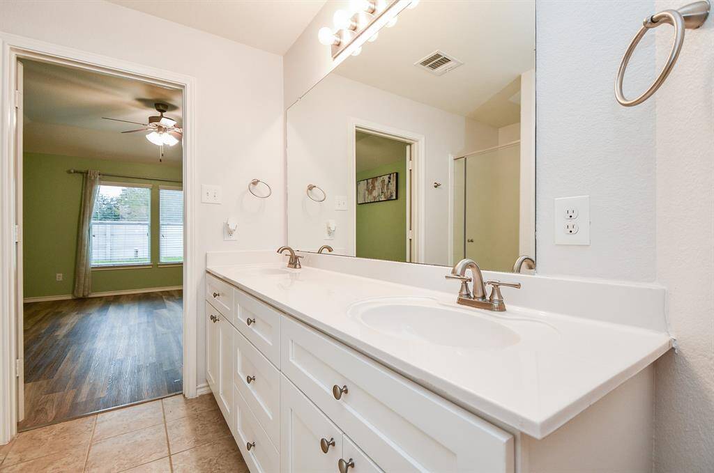 This photo shows a bright bathroom featuring a double sink vanity with ample storage and modern fixtures. It opens into a bedroom with wood flooring, a ceiling fan, and large windows providing natural light.