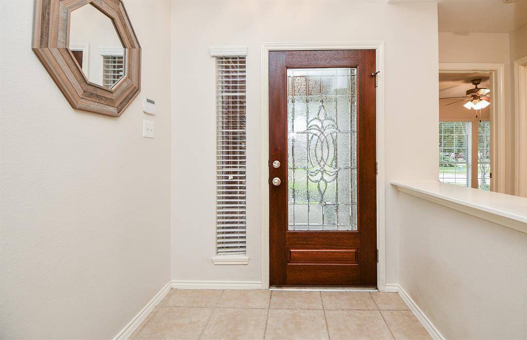 This entryway features a stylish wooden front door with decorative glass, allowing natural light to brighten the space. It includes a side window with blinds and opens to a well-lit interior with tiled flooring.