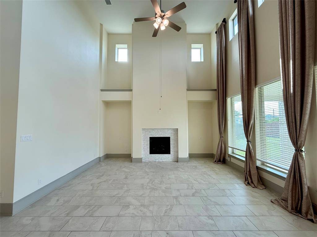 Spacious living room with high ceilings, large windows for natural light, and a central fireplace. Features tile flooring and neutral tones, creating an open and airy atmosphere.