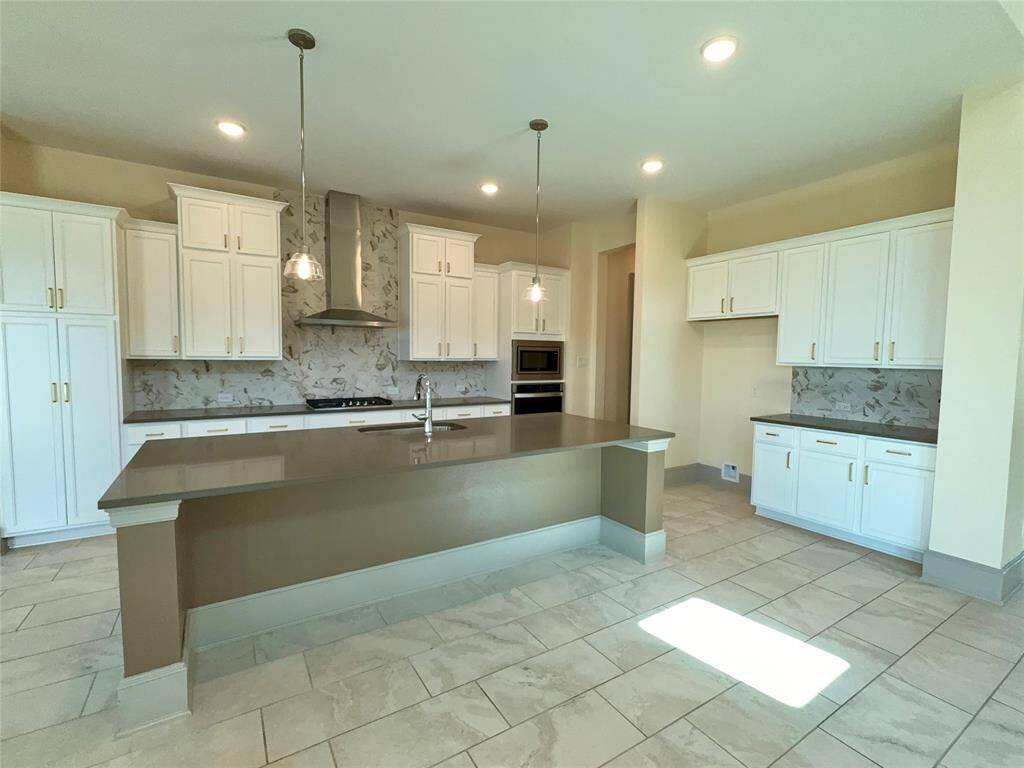This kitchen features modern white cabinetry, a large island with a sleek countertop, stainless steel appliances, and elegant pendant lighting. The backsplash and tile flooring add a stylish touch, creating a bright and inviting space.