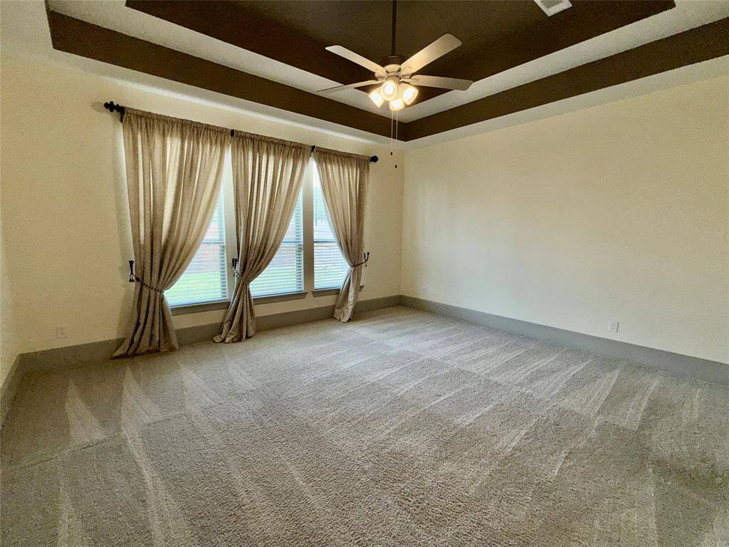 Spacious owner's suite bedroom with carpeted flooring and a ceiling fan. It features large windows with curtains, leading to a bathroom with a visible bathtub. The room has neutral color walls and trim.
