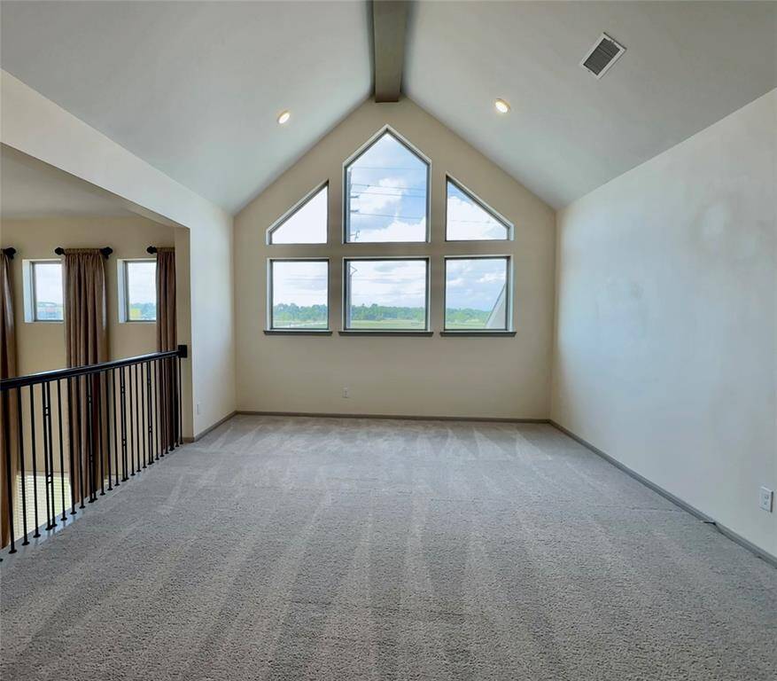 This spacious game room features a vaulted ceiling with recessed lighting and a large triangular window offering abundant natural light and scenic views. The neutral carpet and walls create a versatile space, complemented by a stylish black railing overlooking an open area below.