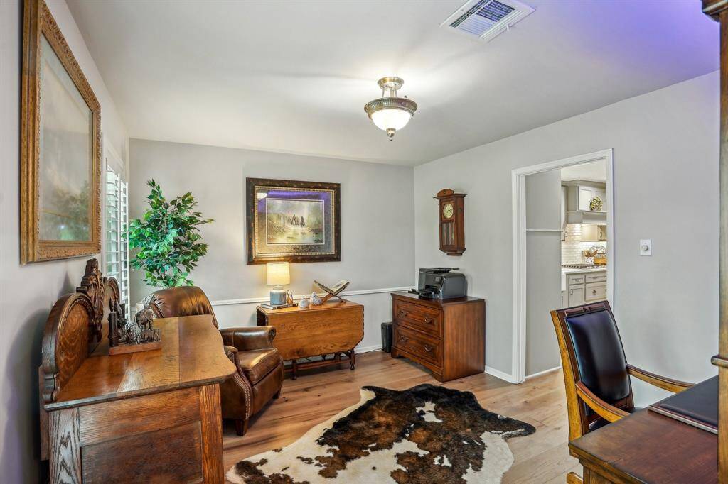 This cozy home office features elegant wooden furniture, a cowhide rug, and soft lighting. It was originally designed as the space for a formal dining area, and it opens into a well-lit kitchen area.