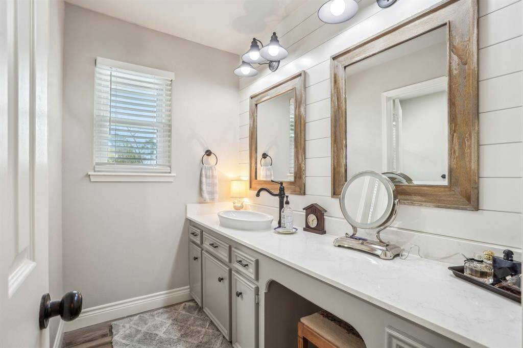 This bathroom features a bright and inviting space with a vanity, rustic wood-framed mirrors, and modern light fixtures. The shiplap wall adds a touch of charm, and a window provides natural light.
