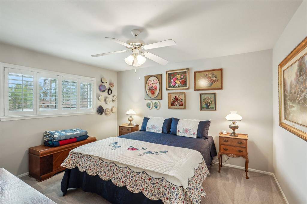 Bedroom featuring a large window with shutters, a ceiling fan. The photo includes a comfortable bed with a quilted cover, bedside tables with lamps, and a wooden storage bench to indicate the spaciousness of this room.