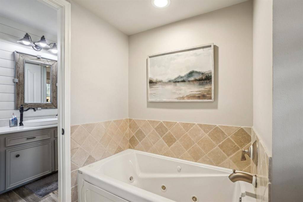 This bathroom features a cozy jetted tub with tile surround, complemented by calming artwork above. Adjacent is a vanity with shiplap wall and a framed mirror, providing a rustic touch.