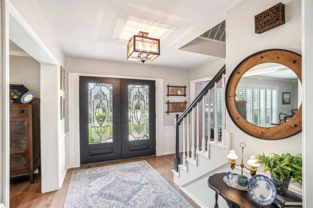 This inviting entryway features elegant double doors with decorative glass, a classic staircase, and a stylish chandelier. A large round mirror and tasteful decor add warmth, while natural light brightens the space.