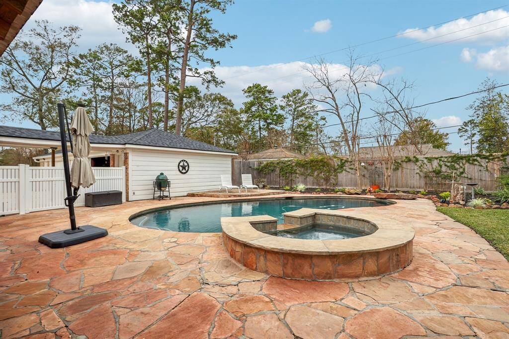 A spacious backyard features a stone patio with an inviting pool and hot tub. The area is surrounded by a wooden fence and lush greenery, creating a private oasis. A detached garage and a patio umbrella add functionality and charm.