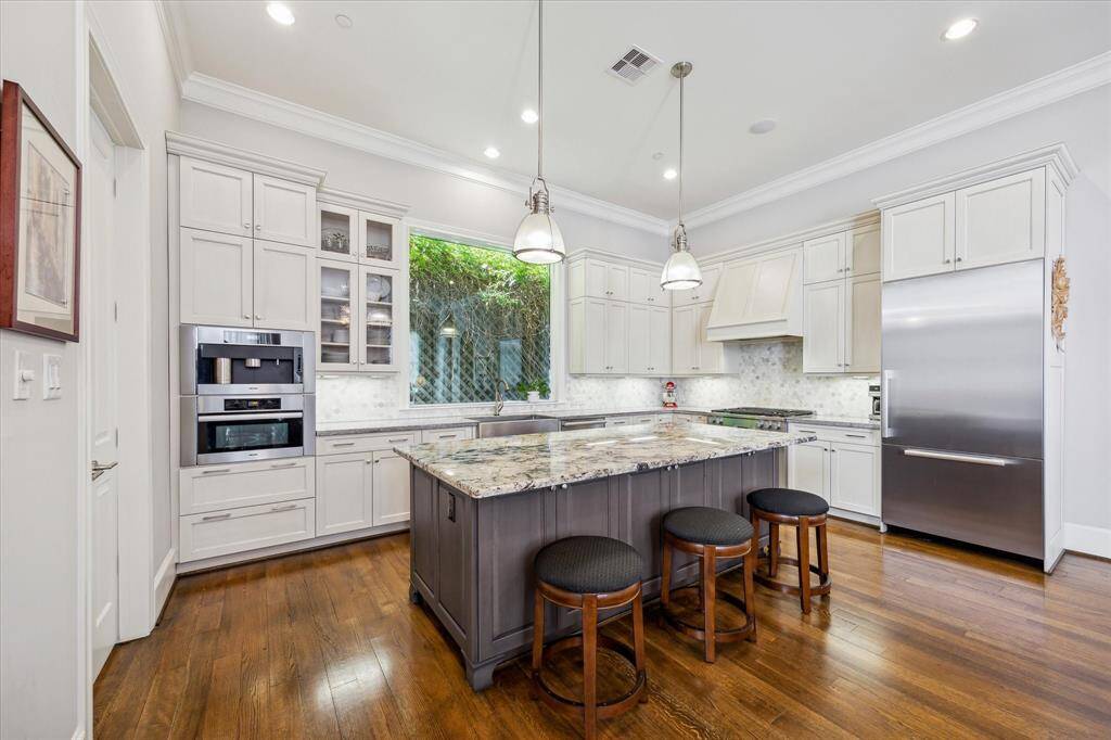 This kitchen features elegant white cabinetry, a spacious granite island with barstools, stainless steel appliances, and warm hardwood flooring. Abundant natural light streams through a large window, creating a bright and inviting atmosphere.