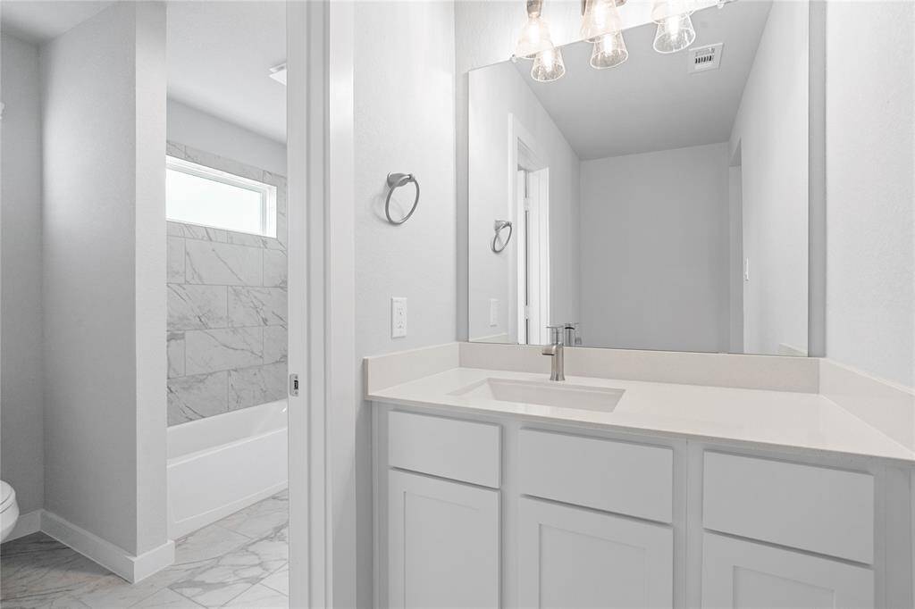 Secondary bath features tile flooring, bath/shower combo with tile surround, stained wood cabinets, beautiful light countertops, mirror, dark, sleek fixtures and modern finishes.