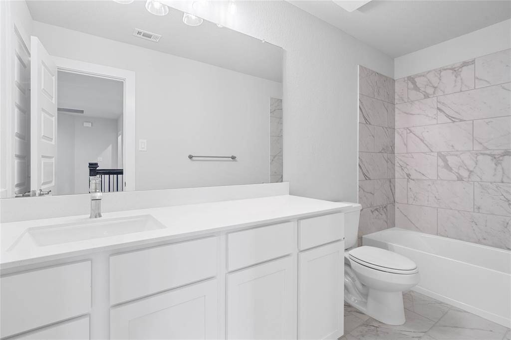 Secondary bath features tile flooring, bath/shower combo with tile surround, stained wood cabinets, beautiful light countertops, mirror, dark, sleek fixtures and modern finishes.