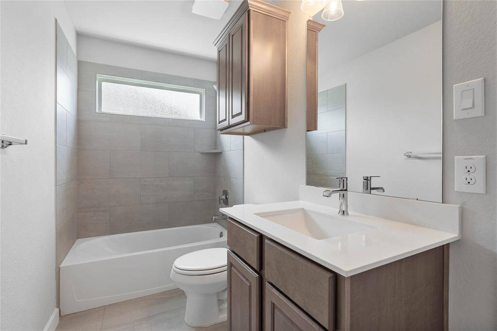 Secondary bath features tile flooring, bath/shower combo with tile surround, stained wood cabinets, beautiful light countertops, mirror, dark, sleek fixtures and modern finishes.