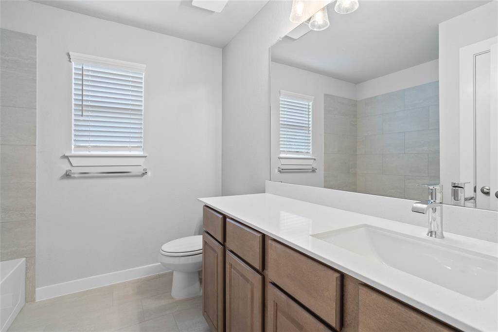 Secondary bath features tile flooring, bath/shower combo with tile surround, stained wood cabinets, beautiful light countertops, mirror, dark, sleek fixtures and modern finishes.