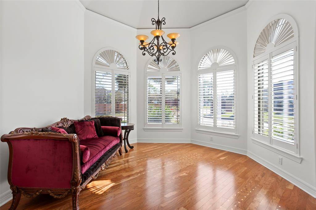 This bright room features elegant hardwood floors, large arched windows with shutters, and a stylish chandelier. The space is enhanced by an ornate burgundy sofa, creating a welcoming and sophisticated atmosphere. Perfect for a sitting area, music room or second home office.