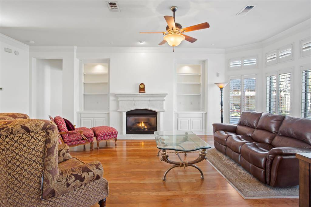 This inviting living room features a cozy fireplace flanked by built-in shelving, a ceiling fan, and rich hardwood floors and is well-lit by large windows with plantation shutters.