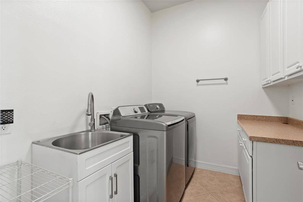 This laundry room features modern appliances, a utility sink, and ample cabinet space. It has a clean, bright design with tiled flooring and a countertop for folding laundry.