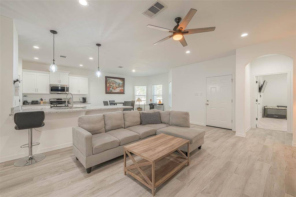 This photo showcases a bright, open-plan living space featuring a modern kitchen with a breakfast bar, a cozy living area with a ceiling fan, and light wood flooring. The room has ample natural light, making it inviting and airy.