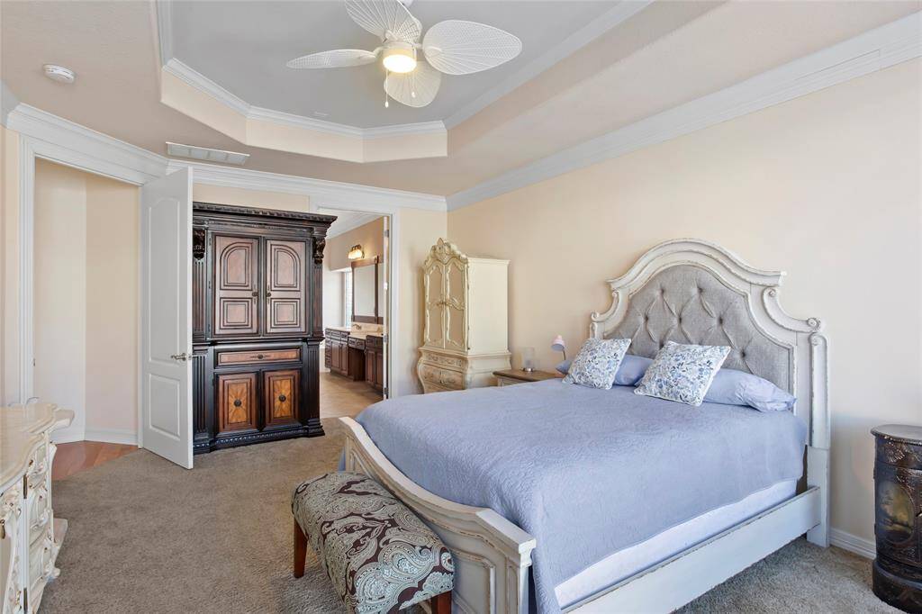 This spacious primary bedroom features a plush carpet, elegant tray ceiling with a fan. The doorway leads to an en-suite bathroom, enhancing convenience and privacy.