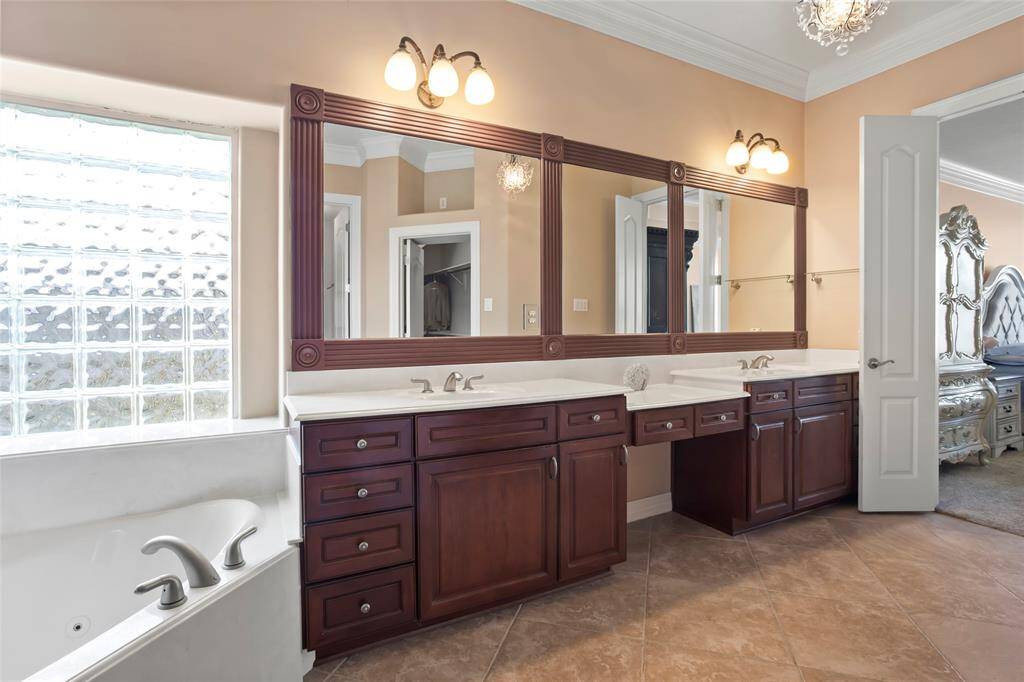 This bathroom features a spacious double vanity with elegant dark wood cabinets and large mirrors. It includes a built-in extra large jetted bathtub with modern fixtures, tile flooring, and ample natural light from a glass block window.